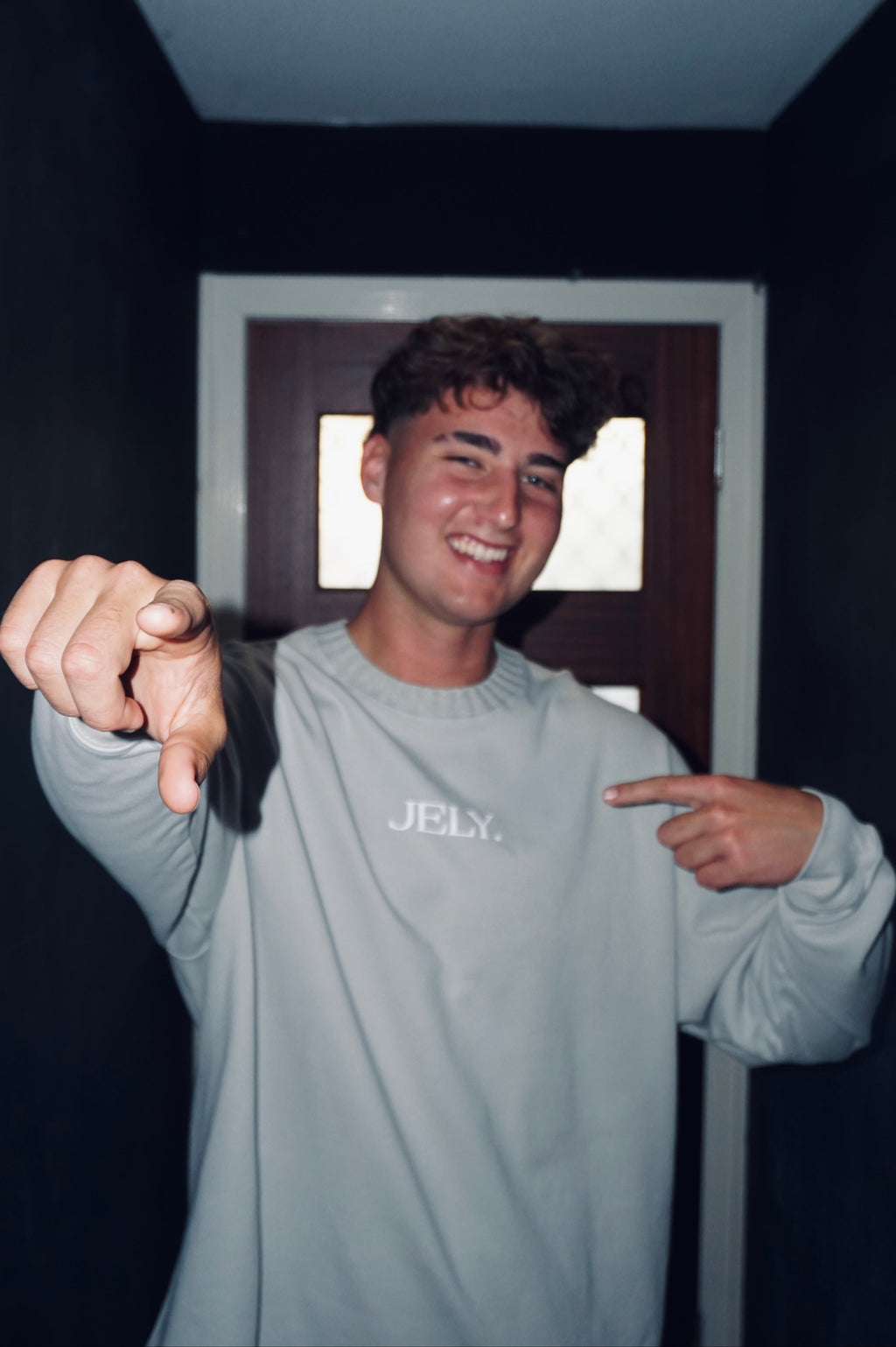 Person wearing a gray 'JELY' shirt pointing forward in a dark room.