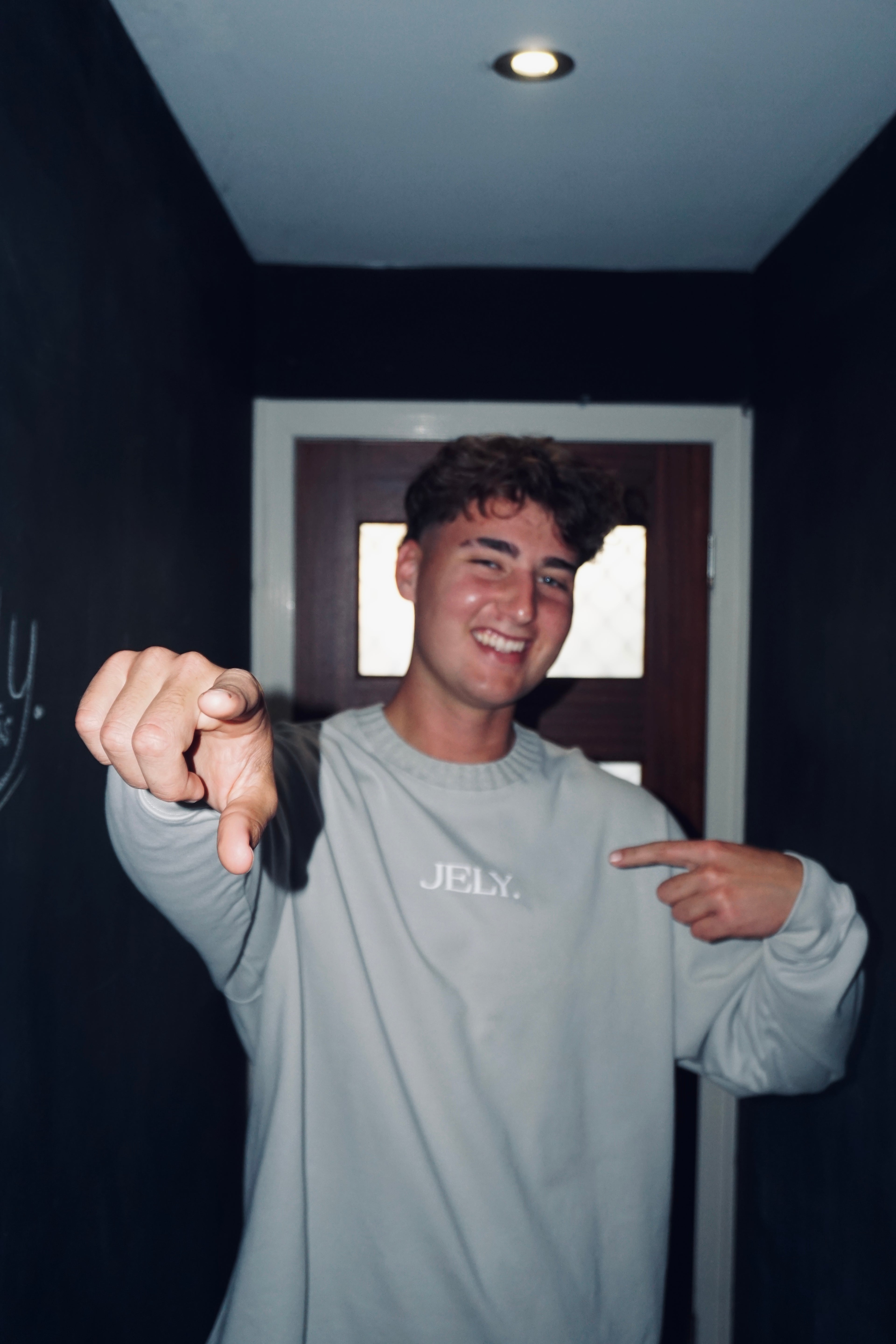 Person wearing a gray 'JELY' shirt pointing forward in a dark room.