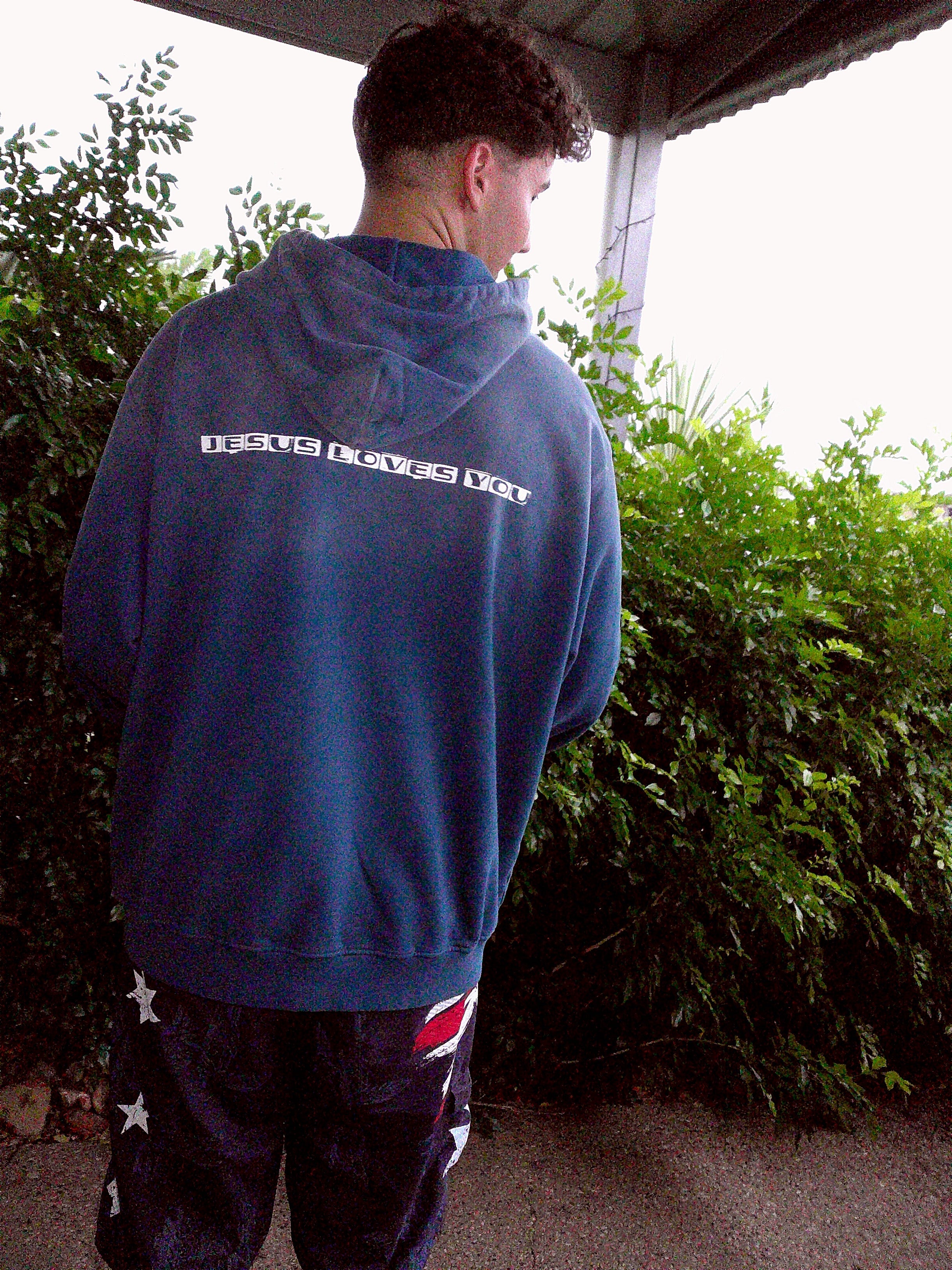 Person wearing a blue sweatshirt with text, standing outdoors near greenery.