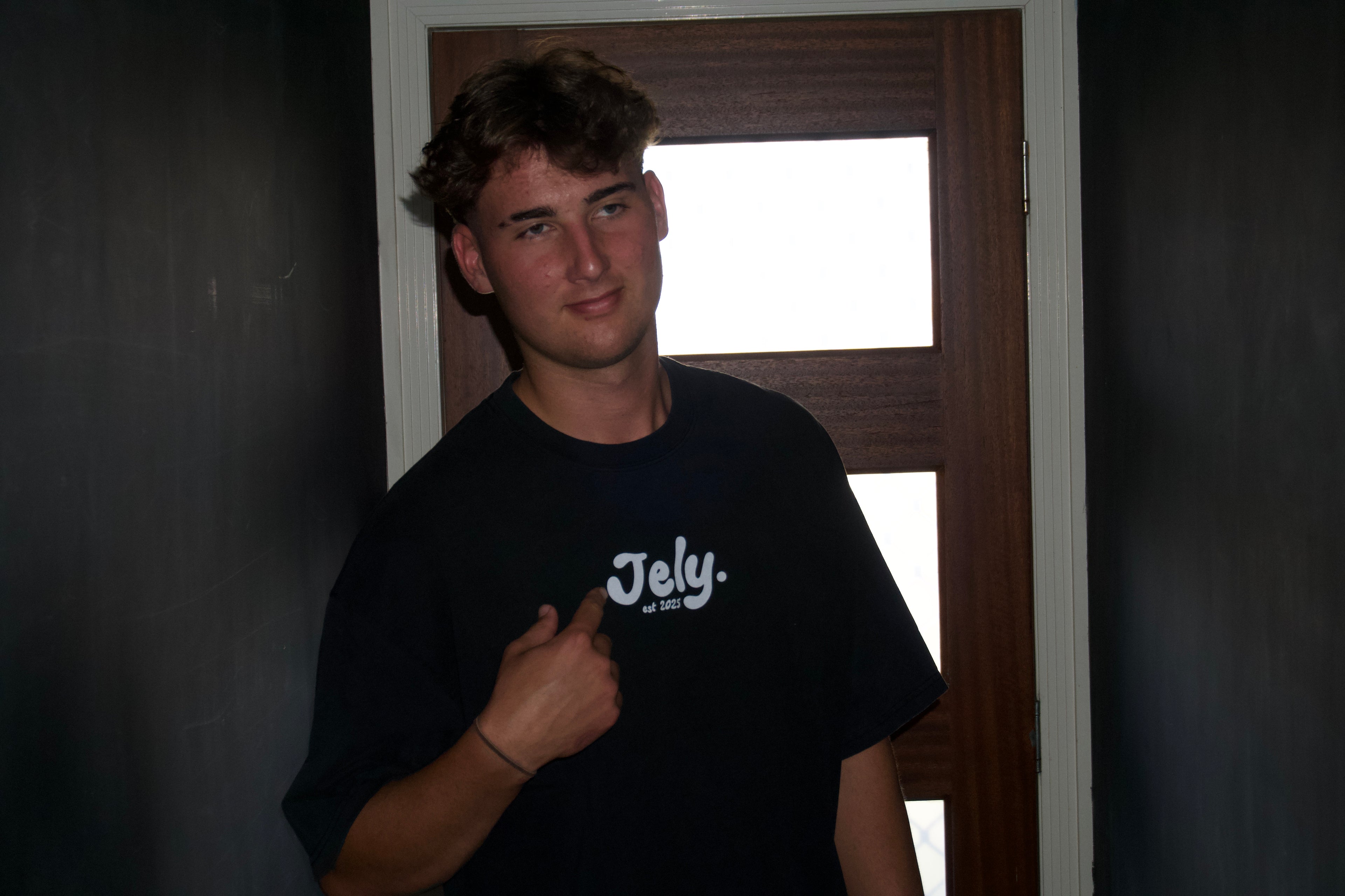 Person wearing a black t-shirt with 'Jely' logo in a dimly lit room.